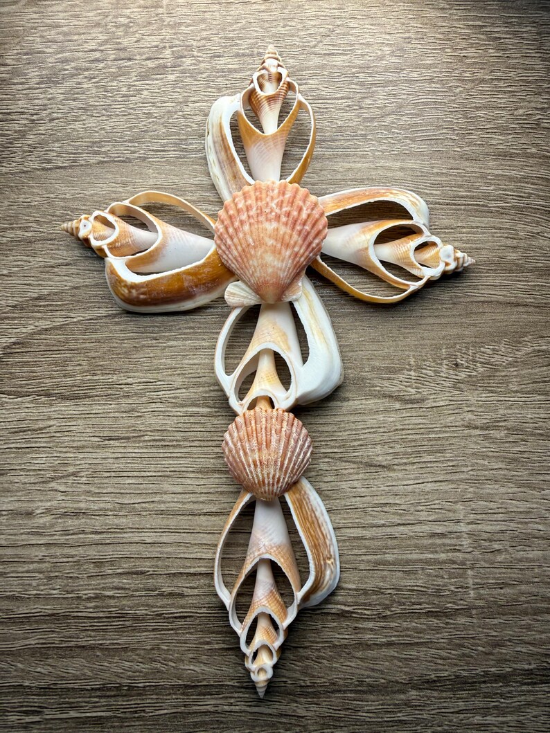 May include: A decorative cross made from various seashells. The cross features a central design of two scallop shells, with other shells forming the arms and base. The shells are primarily cream and tan, with a natural, organic aesthetic.