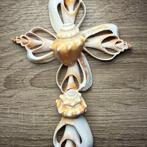 May include: A decorative cross made from various seashells. The cross features a central, spiral-shaped shell in shades of tan and cream, with other shells forming the arms. The background is a wooden surface.