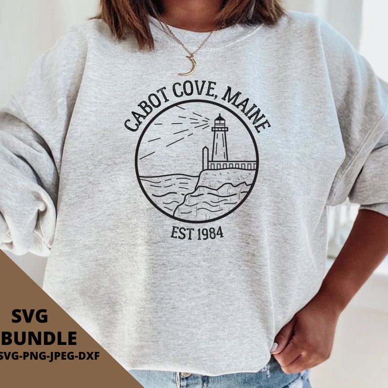 Op de afbeelding: Een grijs sweatshirt met een zwart-wit grafisch ontwerp van een vuurtoren, golven en de tekst "CABOT COVE, MAINE EST 1984".