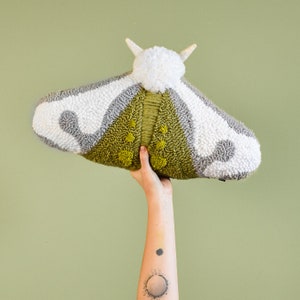 Könnte beinhalten: Ein weißes, graues und grünes Plüsch-Mottenkissen mit einem Bommel auf dem Kopf. Das Kissen wird von einer Hand hochgehalten.