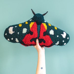 Könnte beinhalten: Eine Hand hält einen grünen, roten und gelben Filzschmetterling mit weißen Akzenten. Der Schmetterling ist mit einer Punch-Needle-Technik hergestellt.