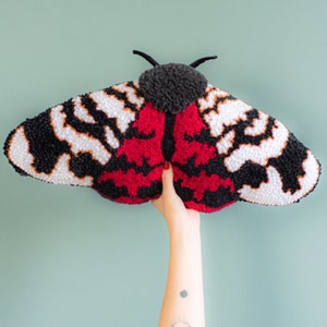 Könnte beinhalten: Ein großer, farbenfroher, handgefertigter Motten-Wandbehang aus Garn. Die Motte hat schwarze, weiße und rote Flügel mit einer flauschigen Textur.
