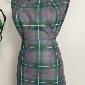 May include: A sleeveless dress featuring a grey, green, and yellow plaid pattern with thin red accent lines. The dress has a square neckline with subtle pleating at the shoulders and a fitted waistline, displayed on a black mannequin form.