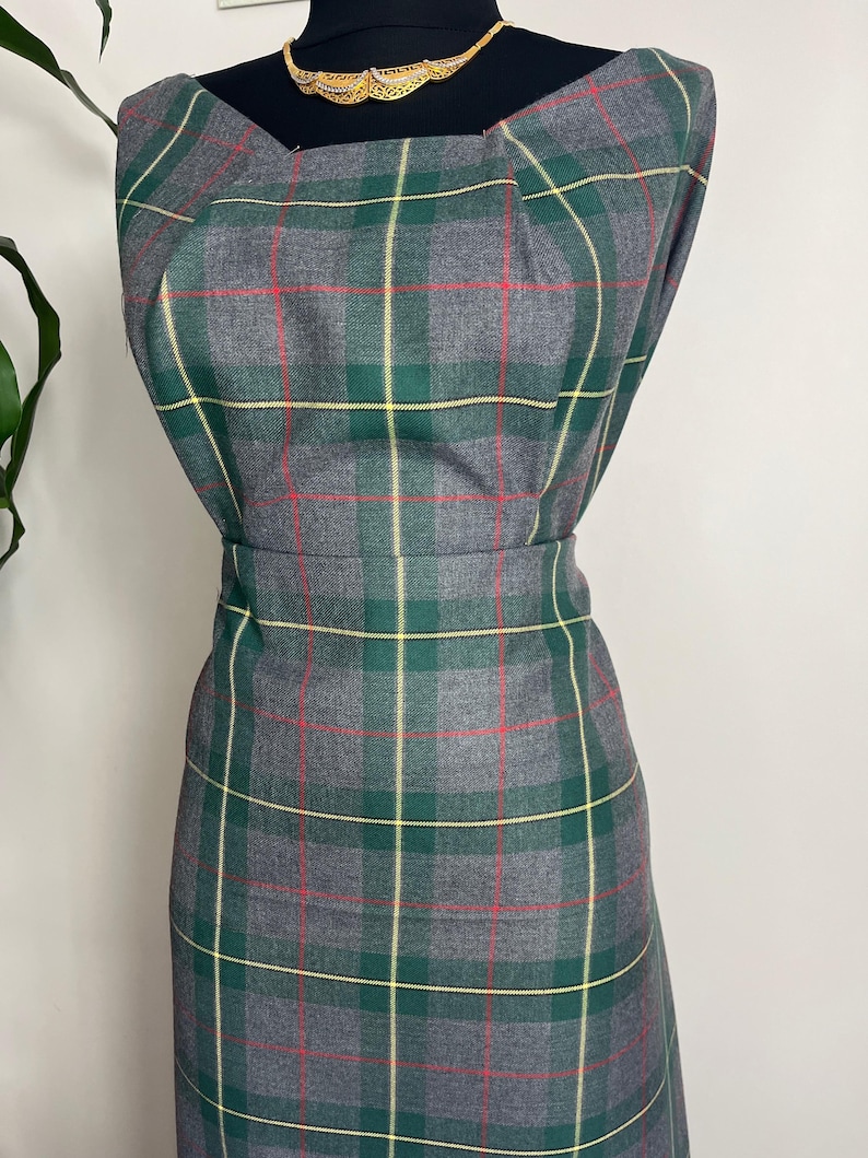 May include: A sleeveless dress featuring a grey, green, and yellow plaid pattern with thin red accent lines. The dress has a square neckline with subtle pleating at the shoulders and a fitted waistline, displayed on a black mannequin form.