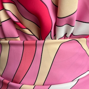 Puede incluir: Primer plano de una tela con un patr&oacute;n vibrante de inspiraci&oacute;n retro. El dise&ntilde;o presenta l&iacute;neas curvas y fluidas en tonos de rosa, rojo, blanco y beige, delineadas en negro. La tela est&aacute; fruncida, creando pliegues y sombras suaves.