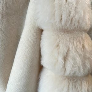 May include: Close-up of a soft, off-white textile with a fluffy, textured accent. The accent appears to be made of multiple stacked, rounded shapes. The fabric has a subtle, woven pattern, creating a cozy and luxurious feel.