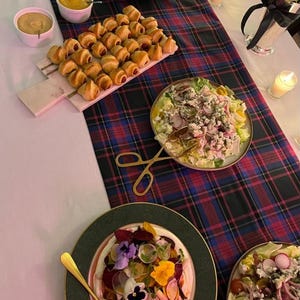 May include: A table setting featuring a rectangular marble serving board with golden-brown crescent rolls, a round plate of salad with radishes and blue cheese, and a plate topped with sliced beets, edible flowers, and a gold serving utensil.