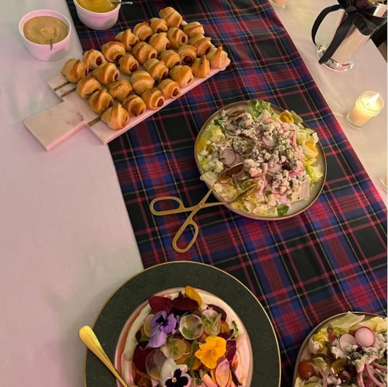 May include: A table setting featuring a rectangular marble serving board with golden-brown crescent rolls, a round plate of salad with radishes and blue cheese, and a plate topped with sliced beets, edible flowers, and a gold serving utensil.