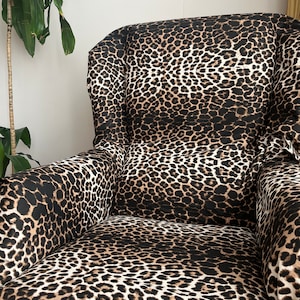 May include: An armchair upholstered in a fabric featuring a repeating leopard print pattern in shades of black, brown, and tan. The chair has a high backrest, integrated side wings, and curved armrests, all covered in the same animal print textile.