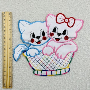 May include: Two embroidered white kittens with pink and blue accents sitting in a colorful basket. The kittens have red bows and black whiskers.