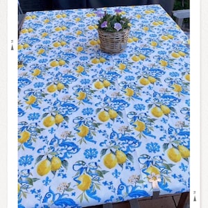 May include: A rectangular tablecloth with a white background and a repeating pattern of yellow lemons and blue floral designs. A small wicker basket with purple flowers sits on the table. The table is made of wood.
