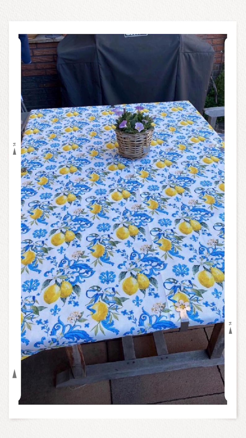 May include: A rectangular tablecloth with a white background and a repeating pattern of yellow lemons and blue floral designs. A small wicker basket with purple flowers sits on the table. The table is made of wood.