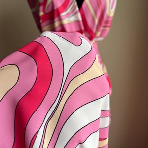 Puede incluir: Una tela vibrante con un estampado abstracto en tonos rosa, blanco y beige. La tela est&aacute; drapeada sobre un maniqu&iacute;, mostrando un dise&ntilde;o fluido. El patr&oacute;n presenta l&iacute;neas curvas, creando una est&eacute;tica retro y psicod&eacute;lica. La tela parece ser un vestido.