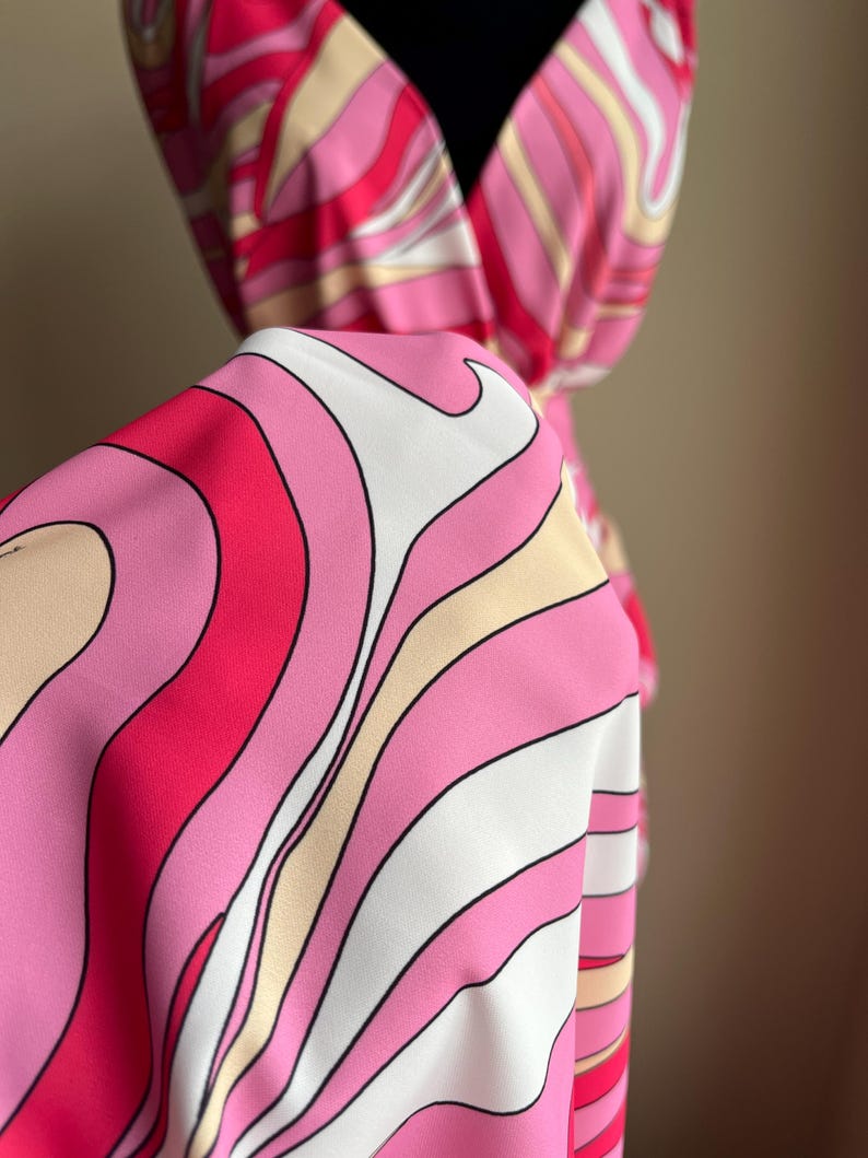 Puede incluir: Una tela vibrante con un estampado abstracto en tonos rosa, blanco y beige. La tela est&aacute; drapeada sobre un maniqu&iacute;, mostrando un dise&ntilde;o fluido. El patr&oacute;n presenta l&iacute;neas curvas, creando una est&eacute;tica retro y psicod&eacute;lica. La tela parece ser un vestido.