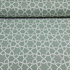 May include: A roll of fabric with a repeating white interlocking pattern on a muted green background. The design is a series of interwoven lines, creating a complex, geometric motif. The fabric appears to be a medium-weight material.