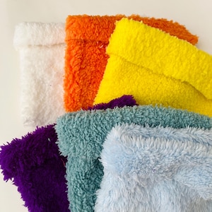 May include: A collection of colorful, fluffy fabric pouches in various shades. The pouches are in white, orange, yellow, purple, teal, and light blue. The fabric has a soft, textured appearance.