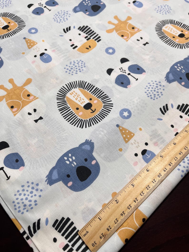 May include: A light blue fabric featuring a repeating pattern of cartoon animals including lions, koalas, pandas, giraffes, and zebras, alongside small stars and dots. A wooden ruler is placed diagonally, showing measurements in inches.