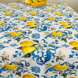 May include: White tablecloth with a repeating pattern of yellow lemons, blue floral accents, and green leaves. A bowl of fruit sits on the table. The tablecloth is made of a durable fabric.
