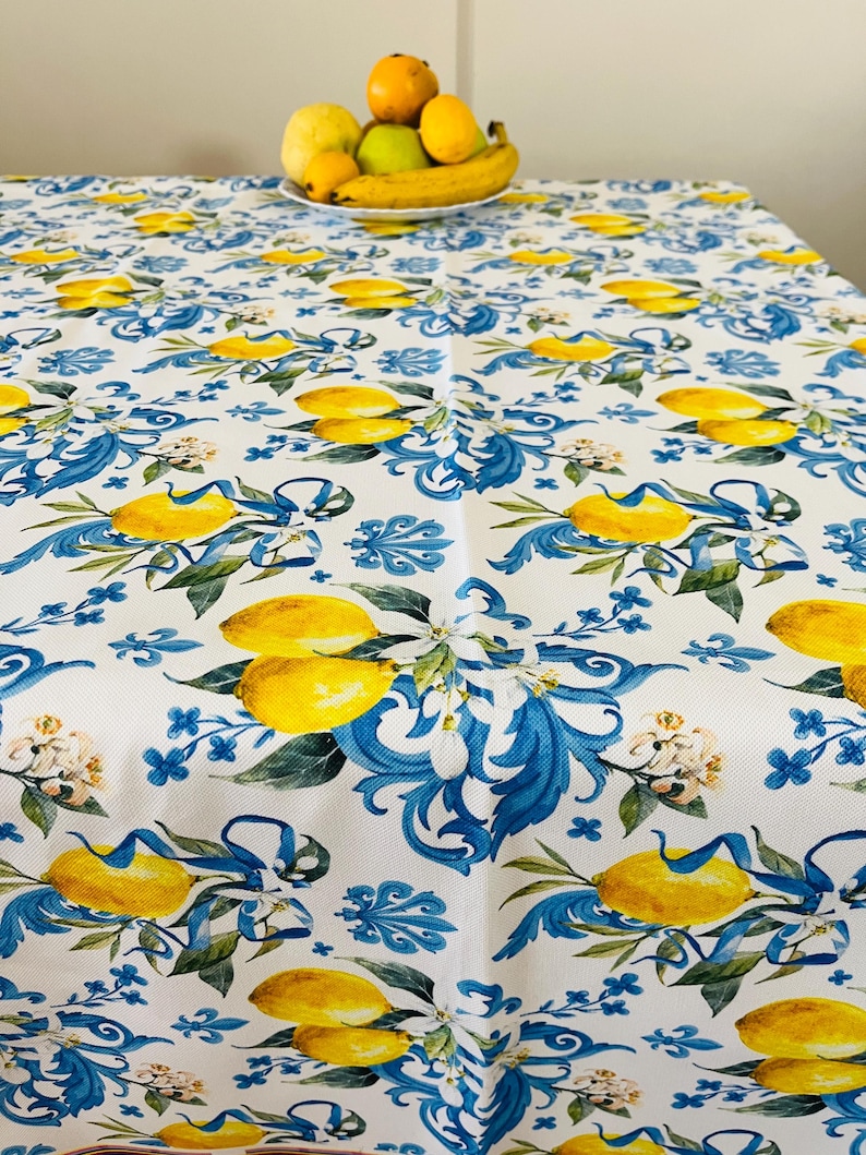 May include: White tablecloth with a repeating pattern of yellow lemons, blue floral accents, and green leaves. A bowl of fruit sits on the table. The tablecloth is made of a durable fabric.