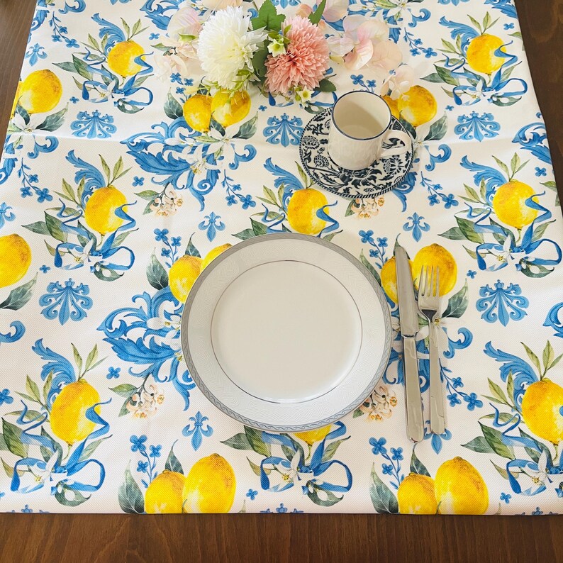 May include: A white tablecloth with a lemon and blue ribbon pattern. A white plate with a silver rim, silverware, and a teacup and saucer are on the table. A bouquet of flowers sits in the center.