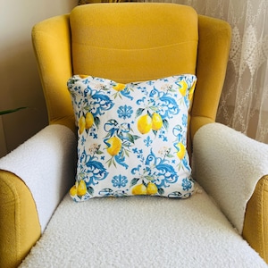 May include: A decorative square pillow with a white background featuring a pattern of yellow lemons, blue floral vines, and white flowers. The pillow is placed on a yellow armchair with a white, fluffy seat cover.