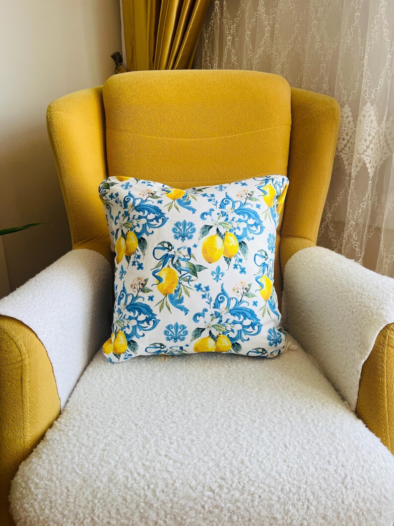 May include: A decorative square pillow with a white background featuring a pattern of yellow lemons, blue floral vines, and white flowers. The pillow is placed on a yellow armchair with a white, fluffy seat cover.