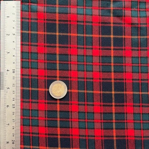 May include: A close-up of a red, green, and navy blue tartan fabric. The fabric features a traditional plaid design with intersecting lines. A silver ruler with inch measurements is on the left. A coin is on the fabric.