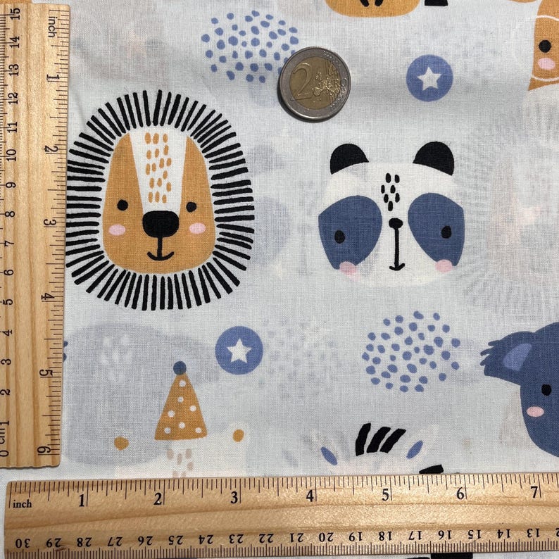 May include: A light gray fabric featuring a repeating pattern of animal faces including a lion, panda, and koala, accented with blue stars and dots. A wooden ruler on the left shows a length of 15 inches, and a coin is placed for scale.