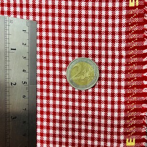 May include: A close-up of red and white checkered fabric with a 2 Euro coin and a ruler. The fabric has a classic gingham pattern. The coin is approximately 1 inch in diameter. The ruler shows measurements in millimeters.