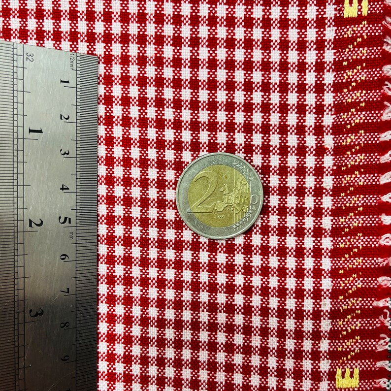 May include: A close-up of red and white checkered fabric with a 2 Euro coin and a ruler. The fabric has a classic gingham pattern. The coin is approximately 1 inch in diameter. The ruler shows measurements in millimeters.