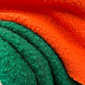 May include: Close-up of two layers of textured fabric. The top layer is a vibrant orange, and the bottom layer is a deep green. The fabric has a soft, fuzzy texture, with visible curls and a slight sheen. The folds of the fabric create shadows and depth.