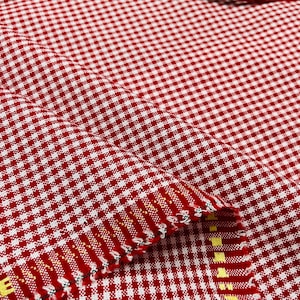 May include: Close-up of red and white checkered fabric. The fabric has a classic gingham pattern, with small, evenly spaced squares. The fabric is folded, showing the texture and weave of the material. The edges are finished with a decorative stitch.