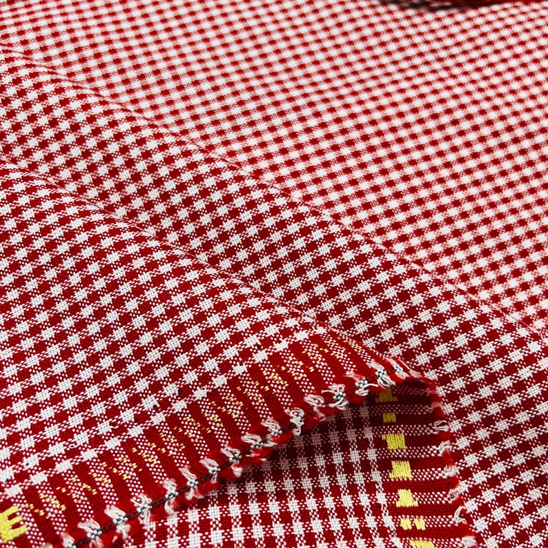 May include: Close-up of red and white checkered fabric. The fabric has a classic gingham pattern, with small, evenly spaced squares. The fabric is folded, showing the texture and weave of the material. The edges are finished with a decorative stitch.