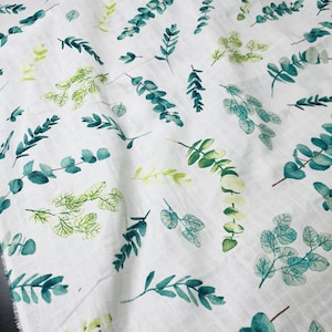 May include: A white fabric featuring a repeating pattern of watercolour-style eucalyptus leaves and branches in various shades of green, ranging from light lime to deep teal, printed across the textured, lightweight, semi-sheer textile surface.