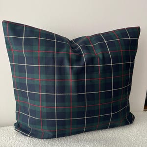 May include: A square throw pillow featuring a classic tartan plaid pattern. The fabric displays a repeating grid of dark navy blue and forest green blocks, accented by thin, intersecting horizontal and vertical lines in white and bright red.