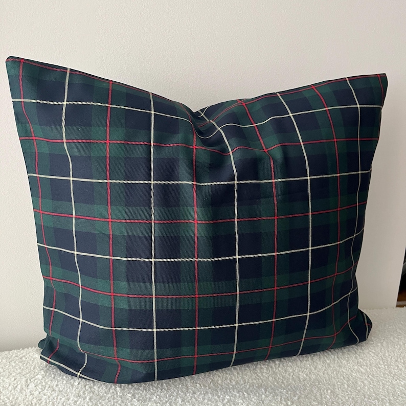 May include: A square throw pillow featuring a classic tartan plaid pattern. The fabric displays a repeating grid of dark navy blue and forest green blocks, accented by thin, intersecting horizontal and vertical lines in white and bright red.