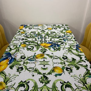 Puede incluir: Mantel rectangular blanco con un dise&ntilde;o de limones y follaje. El patr&oacute;n presenta limones amarillos, hojas verdes y detalles azules. El mantel est&aacute; sobre una mesa rectangular.
