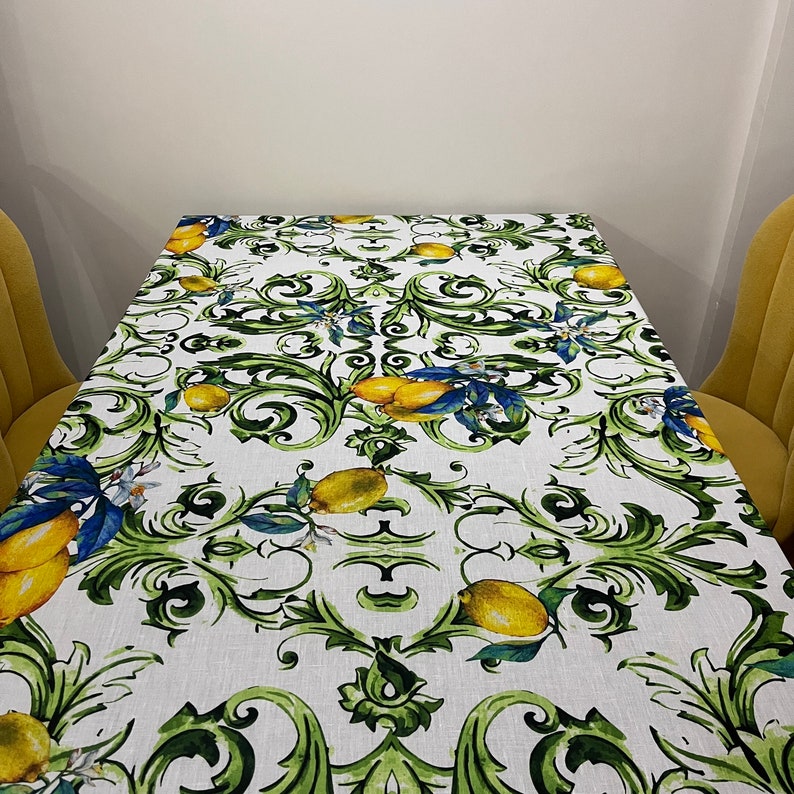 Puede incluir: Mantel rectangular blanco con un dise&ntilde;o de limones y follaje. El patr&oacute;n presenta limones amarillos, hojas verdes y detalles azules. El mantel est&aacute; sobre una mesa rectangular.
