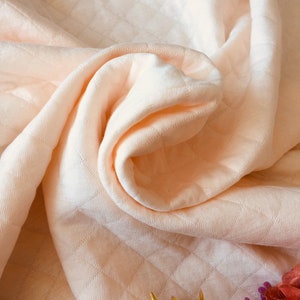 May include: A soft, peach-colored quilted blanket with a subtle diamond pattern. The blanket is folded and draped over a surface, with a small bouquet of flowers in the bottom right corner.