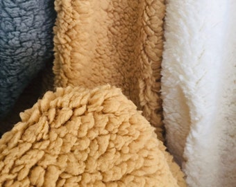 May include: Close-up of three different textures of faux fur fabric. The fabric on the left is a dark grey, the middle fabric is a light brown, and the fabric on the right is white.