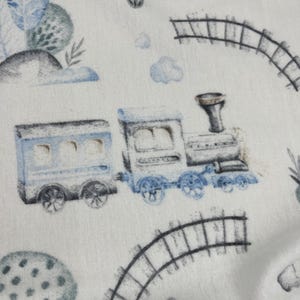 May include: A white fabric with a repeating pattern of blue toy trains, train tracks, and clouds. The trains have black wheels and gray smoke stacks.