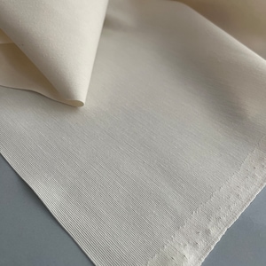 May include: Close-up of a white fabric with a subtle texture and a stitched edge. The fabric is folded over itself, creating a soft, flowing look.