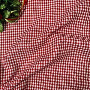 May include: A close-up of a red and white checkered fabric with a slight wave pattern. The fabric is a classic gingham pattern, often used for tablecloths and other home decor items. Green leaves and red and gold ribbons are in the upper left corner.