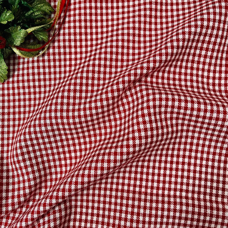 May include: A close-up of a red and white checkered fabric with a slight wave pattern. The fabric is a classic gingham pattern, often used for tablecloths and other home decor items. Green leaves and red and gold ribbons are in the upper left corner.
