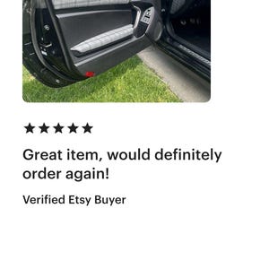 May include: A black car door interior panel featuring grey and white plaid fabric inserts on the upper door trim and the armrest. The door panel is constructed from black plastic with a smooth finish, including a handle and window controls.