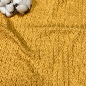 May include: Close-up of a textured, mustard-yellow fabric with a repeating chevron pattern. The fabric has a soft, knitted appearance. A few white cotton bolls are visible in the upper left corner, adding a natural element to the image.