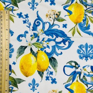 May include: White fabric with a repeating pattern of yellow lemons, blue ribbons, and floral accents. The design includes blue fleur-de-lis and small blue flowers. The fabric is measured with a ruler in inches.