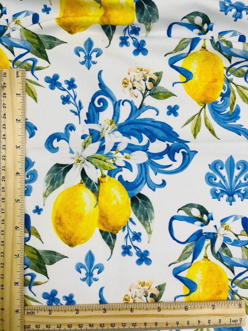 May include: White fabric with a repeating pattern of yellow lemons, blue ribbons, and floral accents. The design includes blue fleur-de-lis and small blue flowers. The fabric is measured with a ruler in inches.