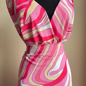 Puede incluir: Un vestido vibrante de estilo retro con un patr&oacute;n arremolinado en tonos de rosa, blanco y beige. El vestido presenta un escote en V y una cintura ajustada, creando una silueta favorecedora. La tela tiene un brillo sedoso, lo que aumenta su atractivo vintage.