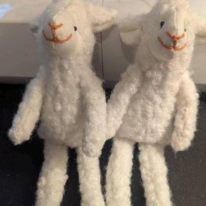 May include: Two handmade white lamb plush toys. Each lamb has a fluffy, textured body, small black eyes, and an orange stitched mouth and nose. The lambs are standing upright with their arms and legs extended.